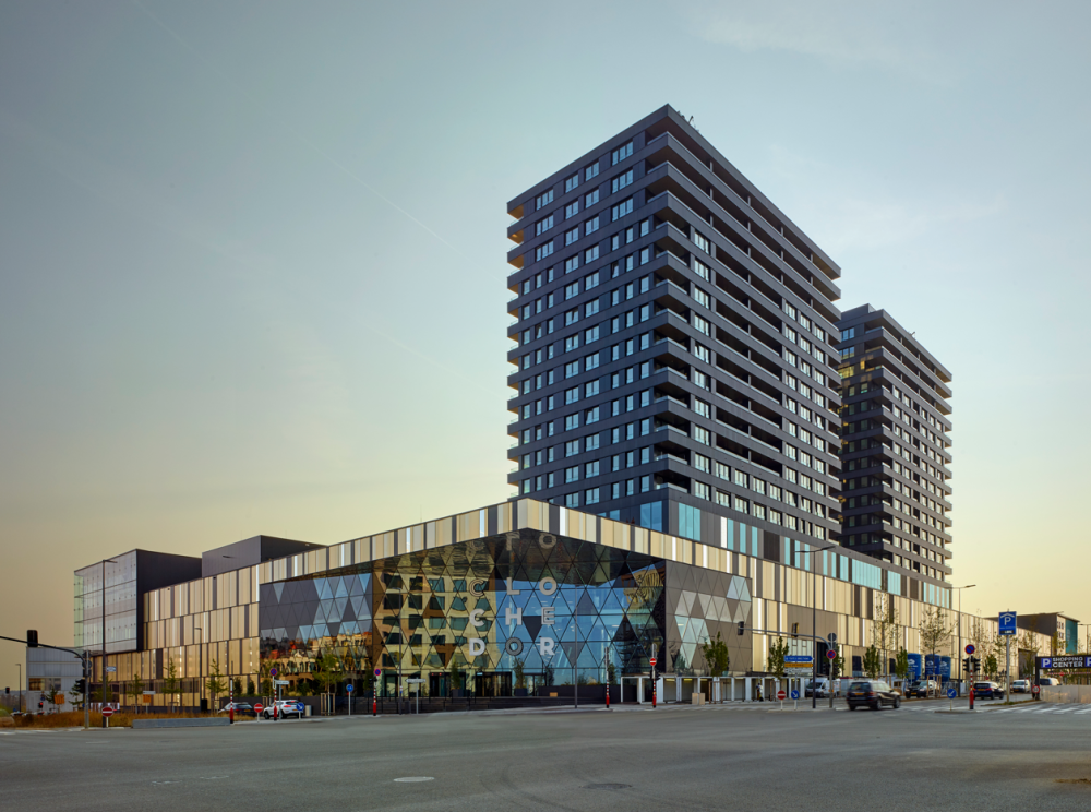 Exterior view of La Cloche d'Or shopping center, a modern building with contemporary architecture, topped by two residential towers. The sky is clear and the esplanade in front of the building is empty.